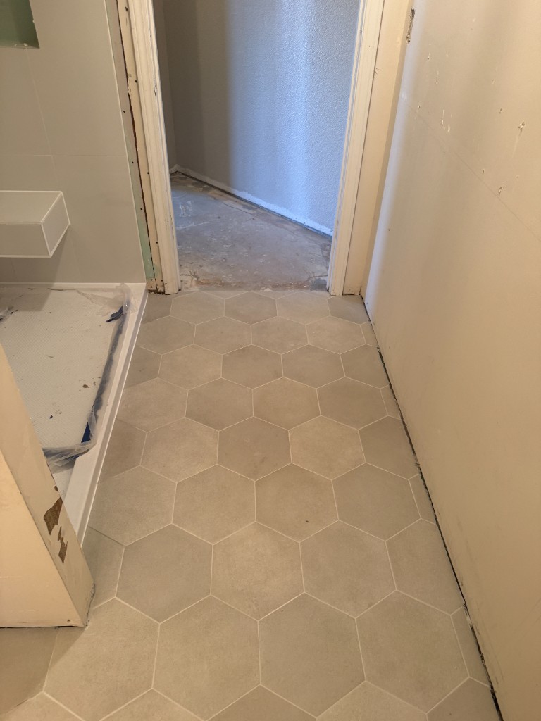Bathroom remodel in progress with large light-gray hexagonal floor tile, shower pan protected, and drywall being finished