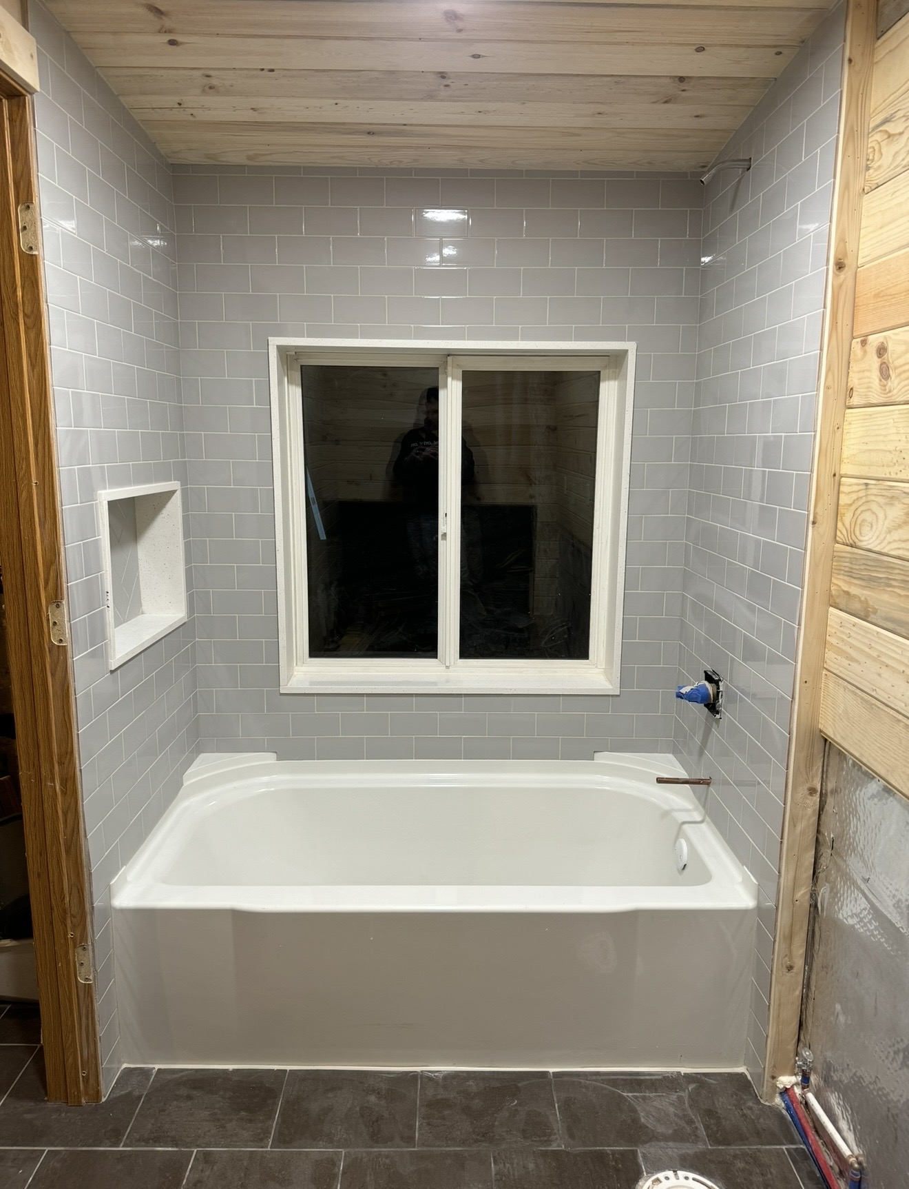 Bathtub alcove with light gray subway tile walls, wood plank ceiling, window, built-in niche, and dark floor tile during renovation