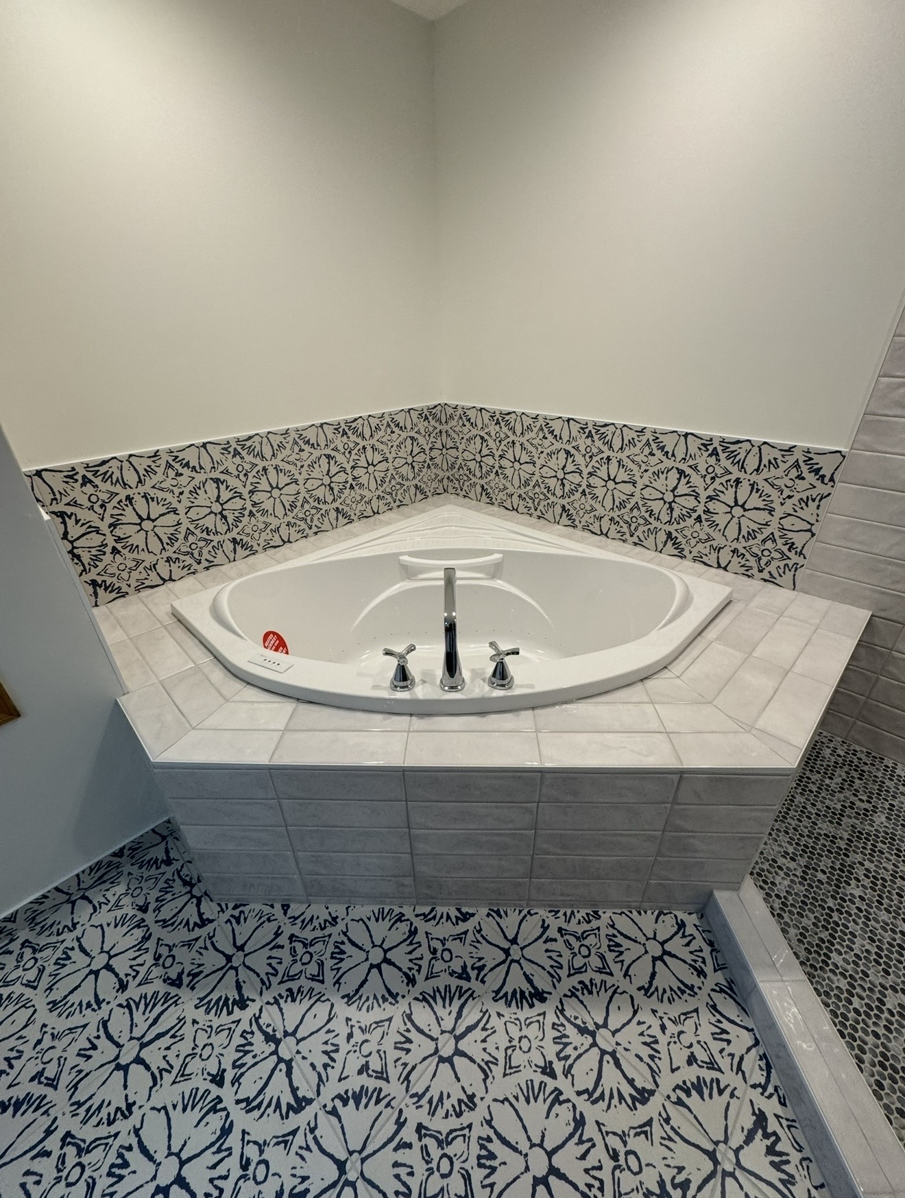 Corner oval drop-in bathtub with light gray tile deck, navy and white floral backsplash, patterned floor tile, and adjacent pebble shower floor