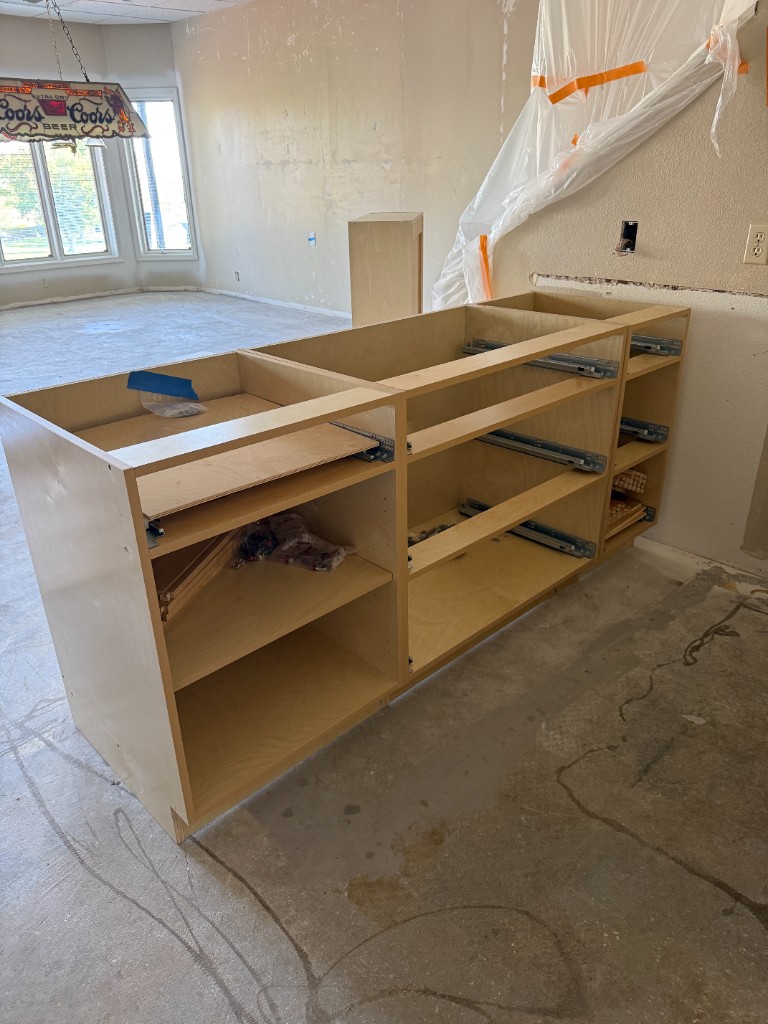 Custom kitchen or bar base cabinets during installation—maple-style boxes on concrete with drawer slides mounted, doors and countertops not yet installed