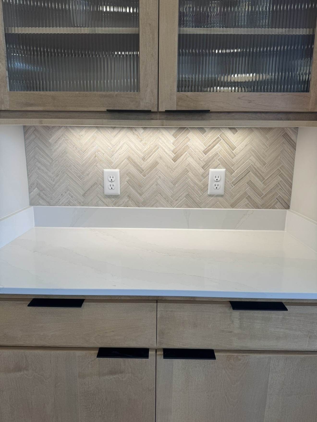 Kitchen or bar cabinetry with light wood finishes, reeded glass upper doors, white quartz countertop, stone splash, and herringbone natural stone tile backsplash with under-cabinet lighting