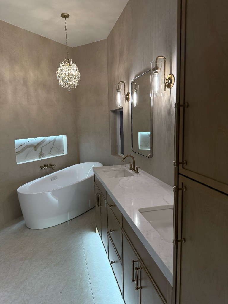 Luxury bathroom with freestanding oval tub, crystal chandelier, lit marble niche, double vanity with marble-look countertop, brass sconces, and warm textured walls