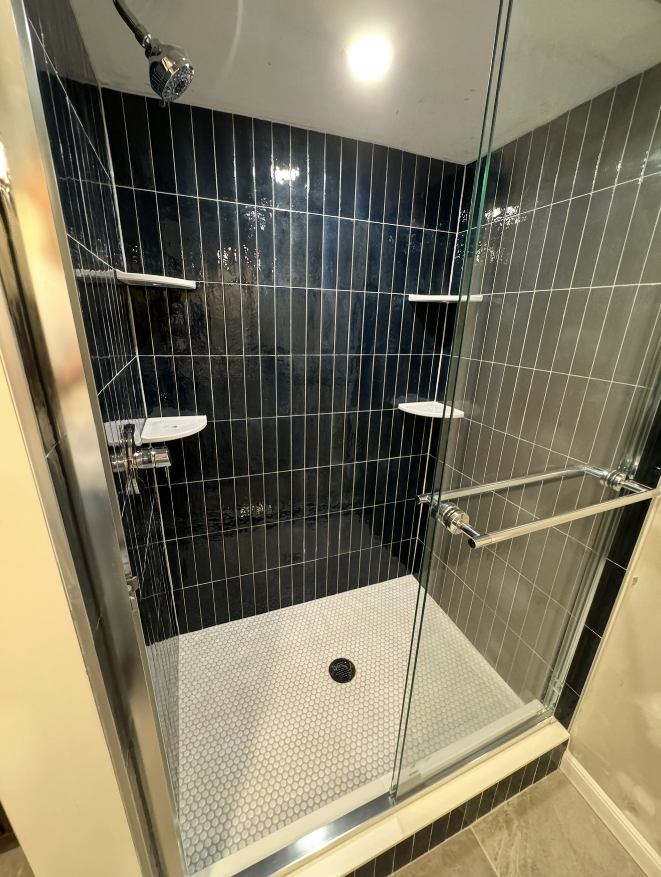 Walk-in shower with vertical navy blue subway tile walls, white penny tile floor, glass enclosure, and chrome fixtures