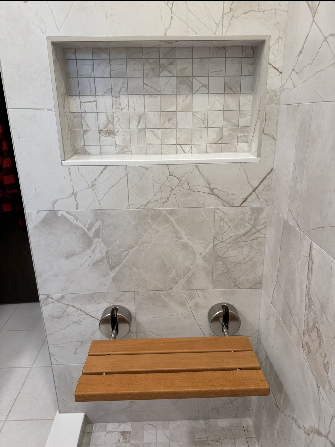 Shower tile niche with mosaic interior and fold-down wood bench mounted on marble-pattern wall with brushed metal brackets