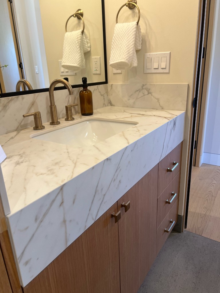 Bathroom vanity with veined marble countertop and backsplash, light wood cabinetry, brushed gold faucet and hardware, and black-framed mirror