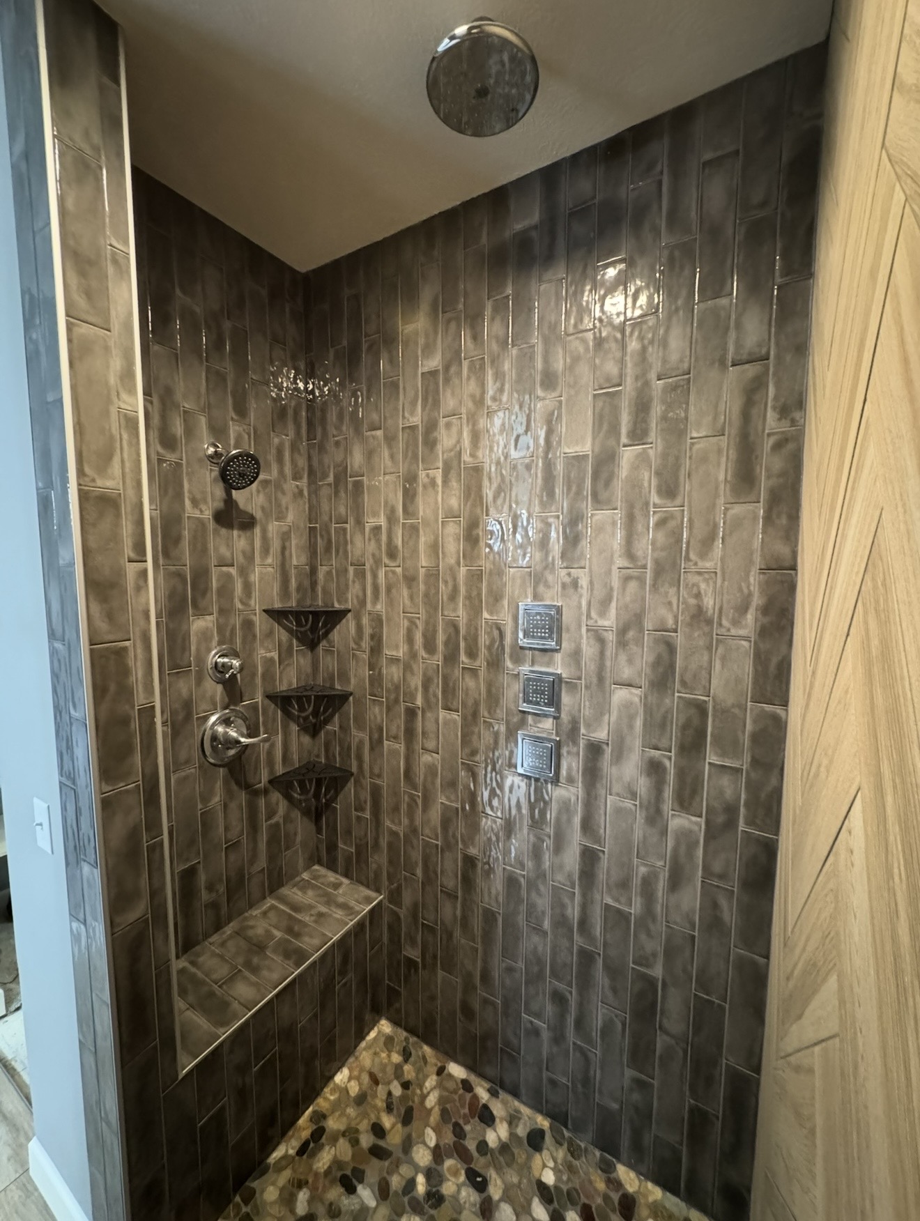 Walk-in shower with dark vertical subway tile, natural pebble floor, tiled bench, corner shelves, body sprayers, rain showerhead, and wood-look herringbone accent wall