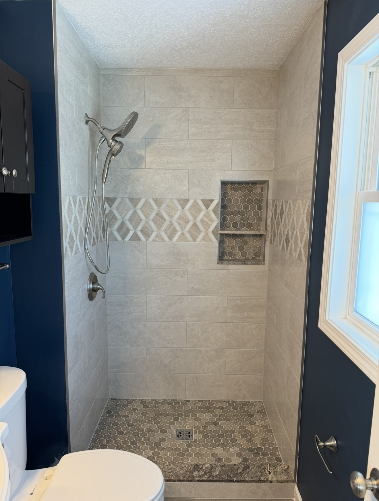 Walk-in shower with stone-look wall tile, decorative accent band, hex tile niche and shower floor, granite curb, and brushed nickel fixtures