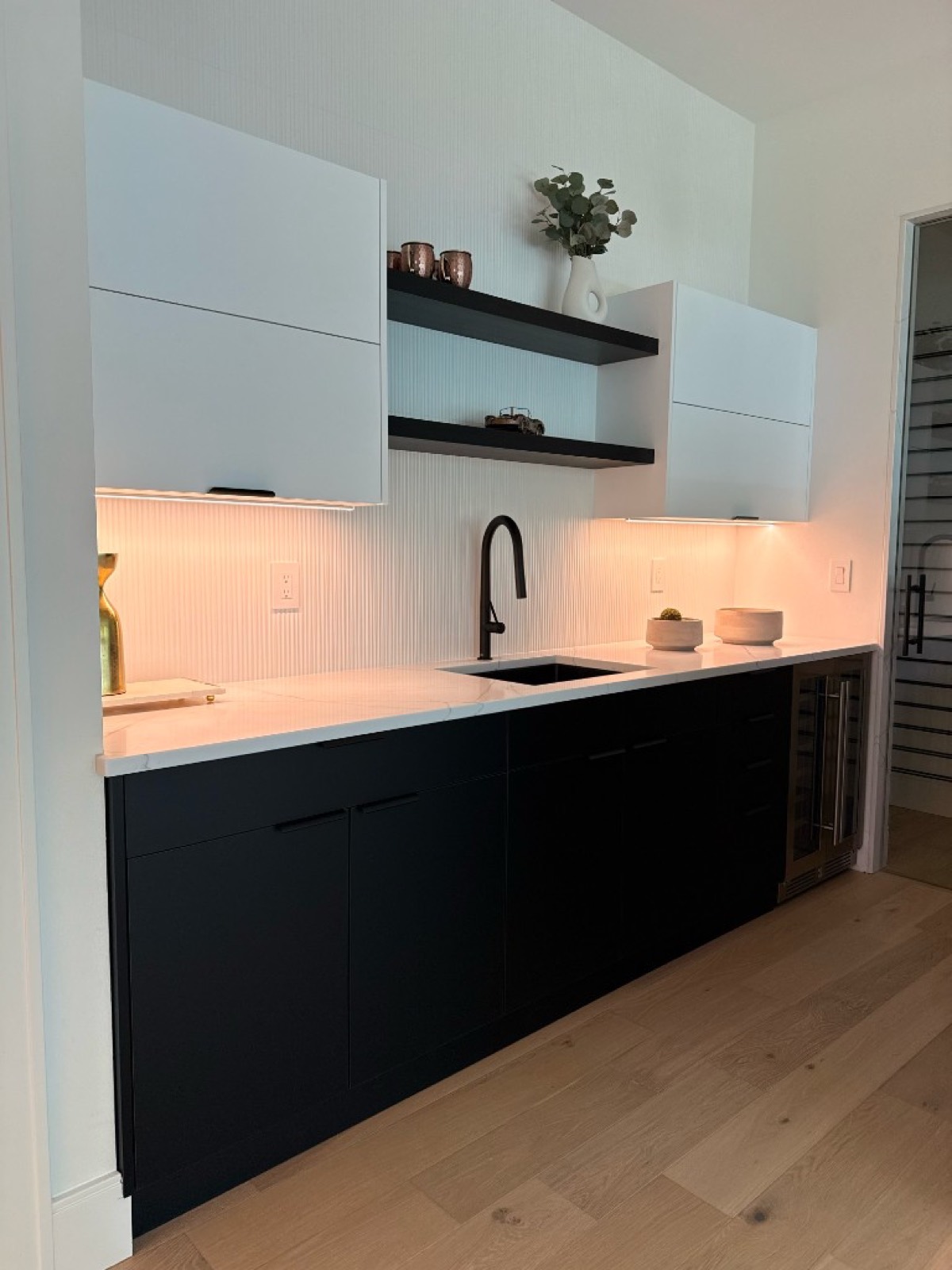 Contemporary wet bar with matte black lower cabinets, white marble countertop, and fluted backsplash